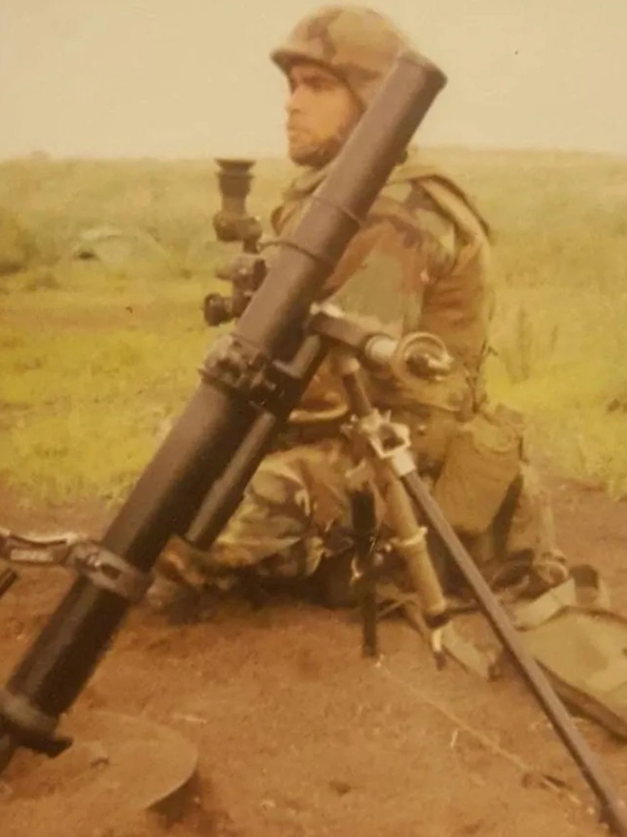 Soldier in camouflage uniform operating a mortar in a grassy field.