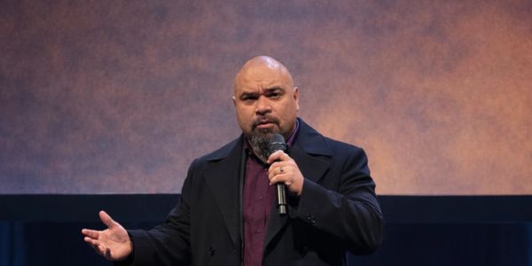 A man speaking into a microphone on stage with a serious expression.