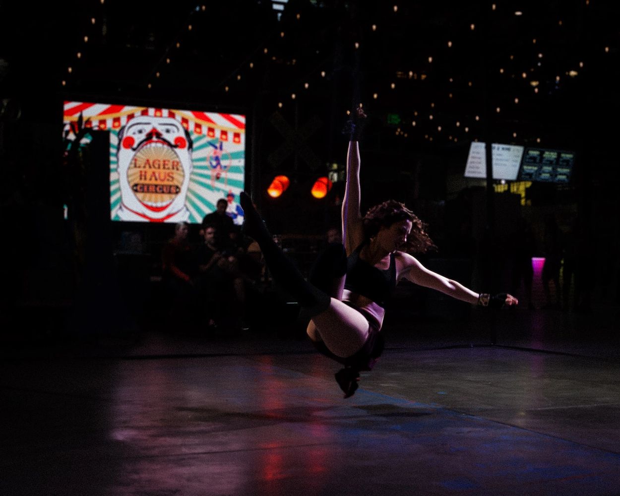 An aerialist looks like she's floating in a dark room. A bright "Lager Haus" logo glows behind her. 