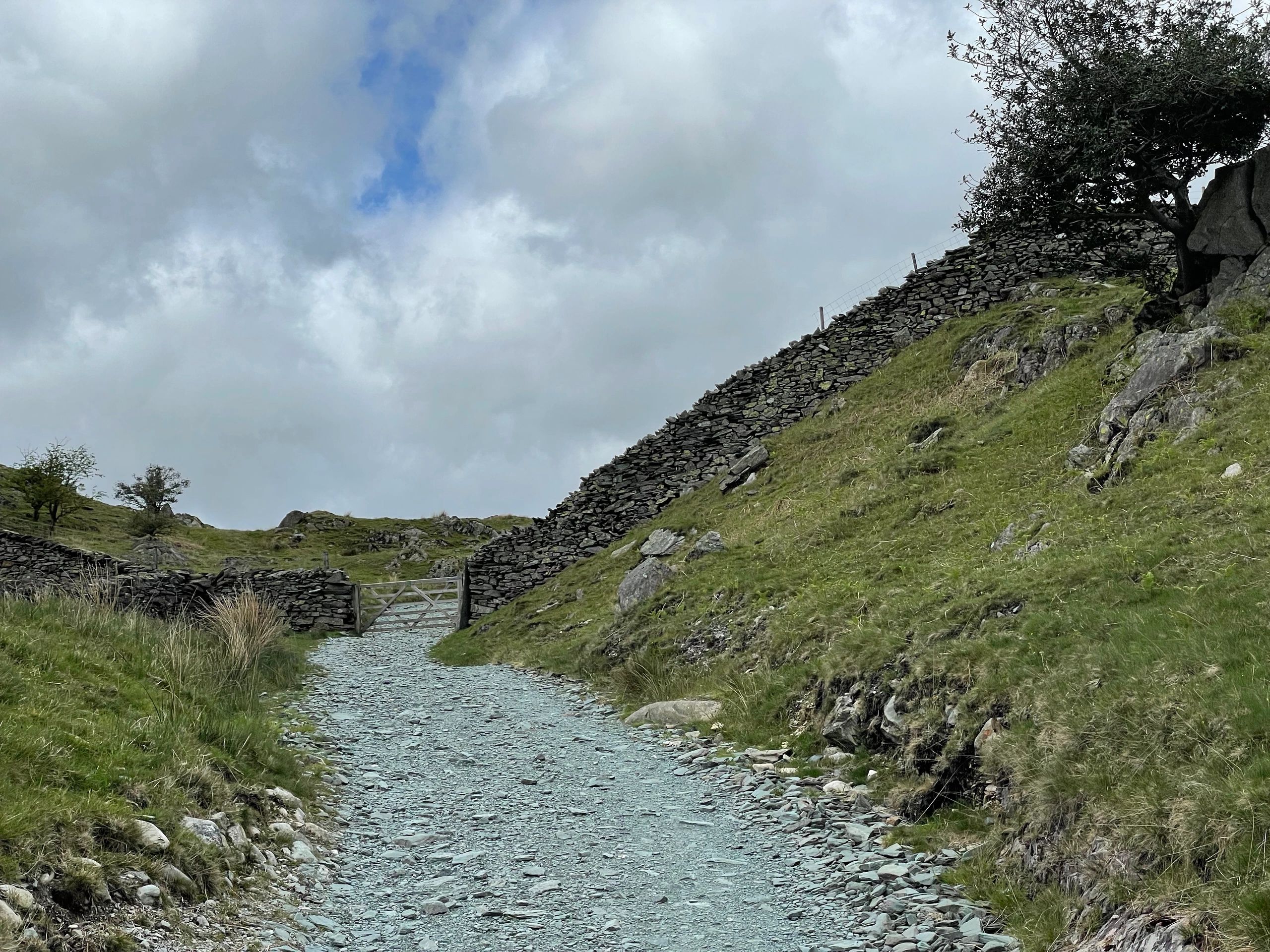 A gravel path leads to a wooden gate with stone walls on a grassy hillside under a cloudy sky.