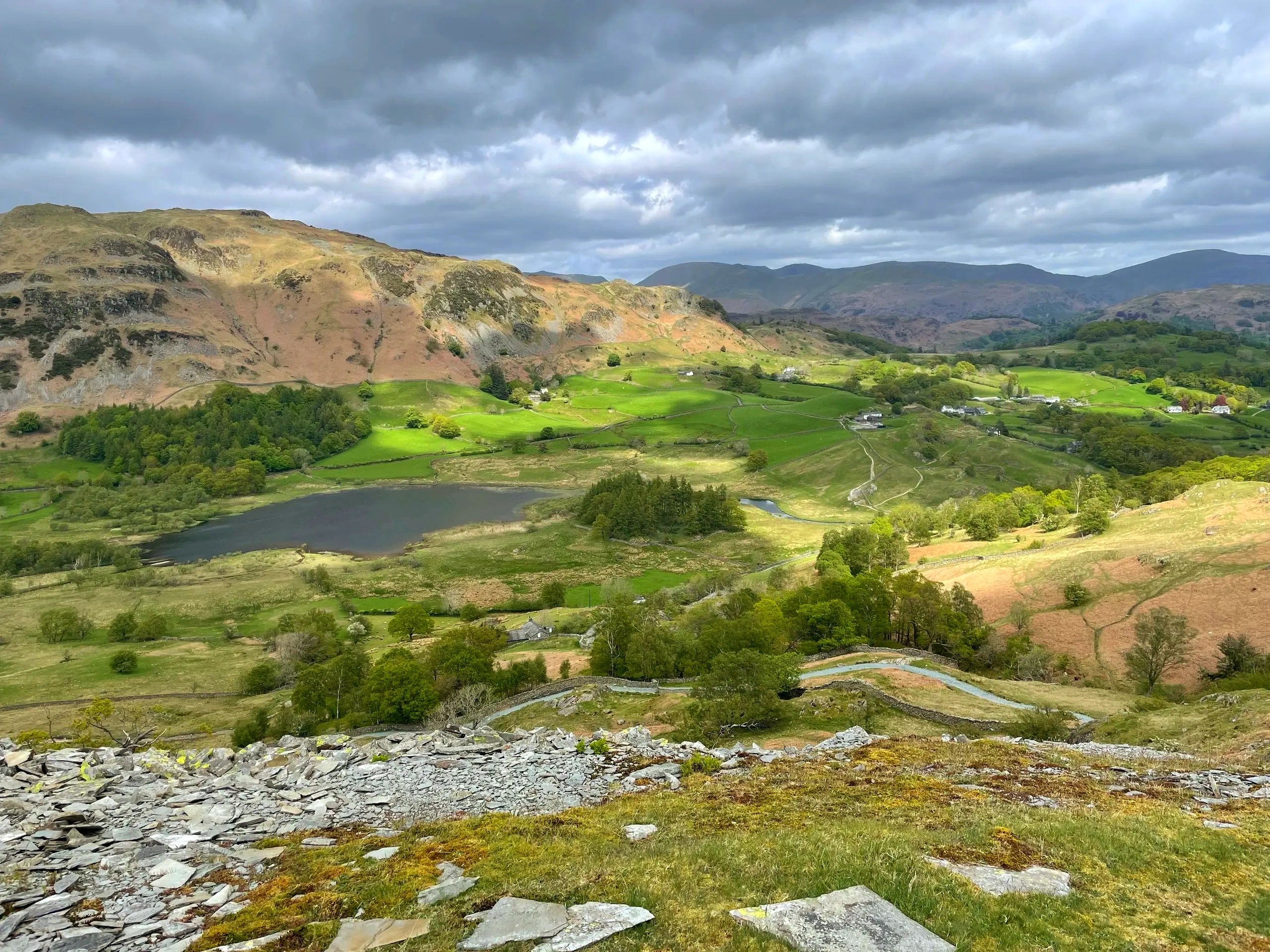Sunlit green hills under a dramatic cloudy sky with a small lake and scattered houses.