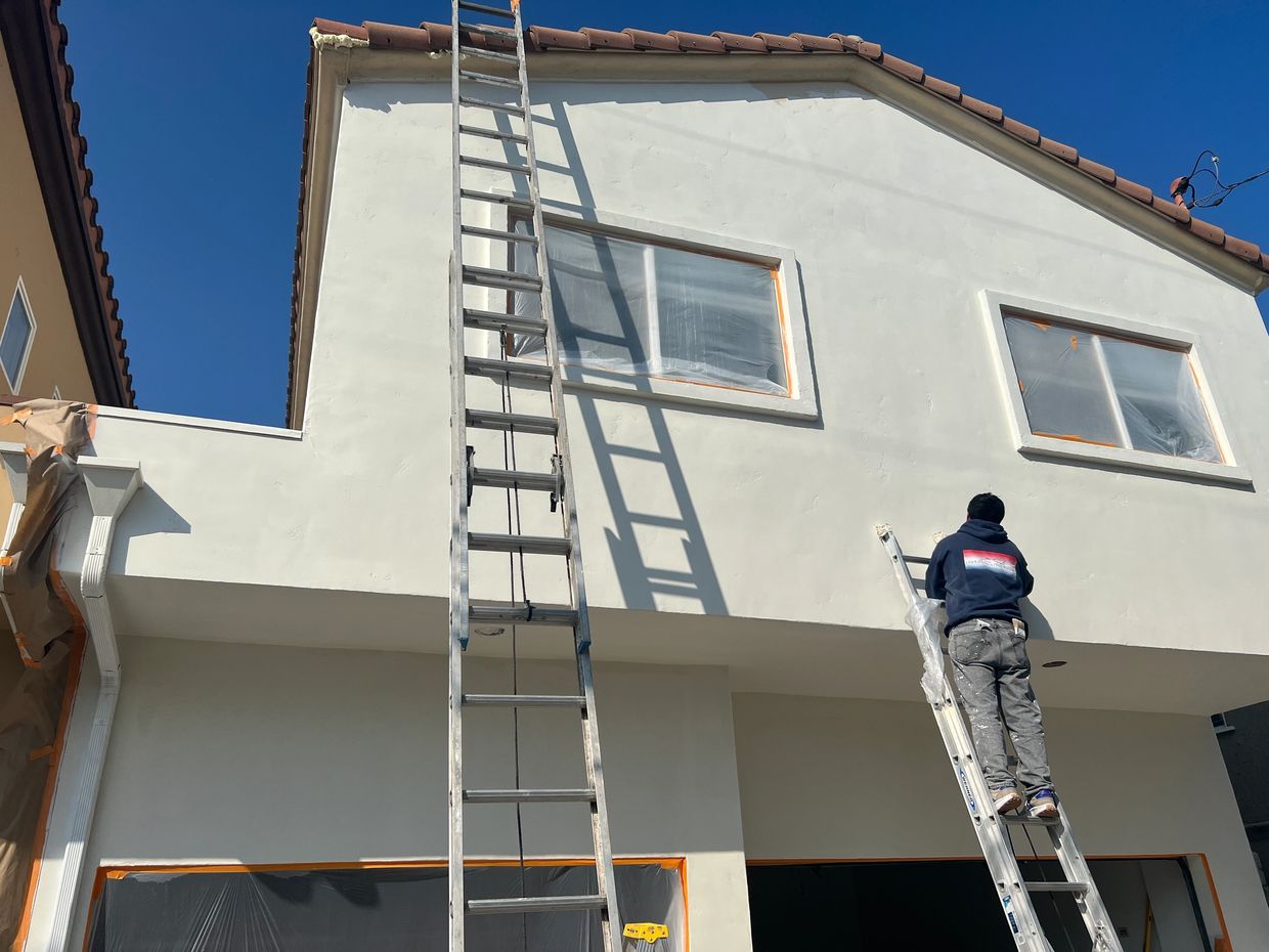 A painter on a ladder working on a white house exterior under clear blue sky.