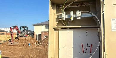 Open electrical panel on construction site with excavator in background under clear blue sky.