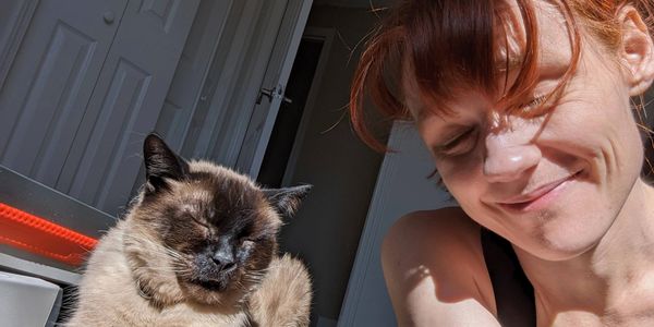 A Siamese cat and a red-haired woman both smile at the camera with their eyes closed