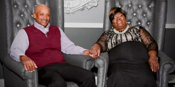 Elderly couple holding hands, sitting on elegant chairs, smiling warmly.