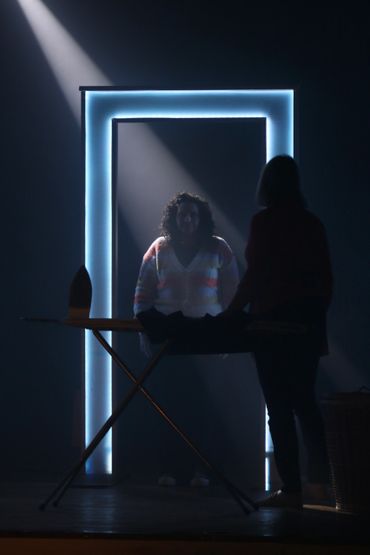 Two people in a dimly lit room with a glowing door frame and ironing board.