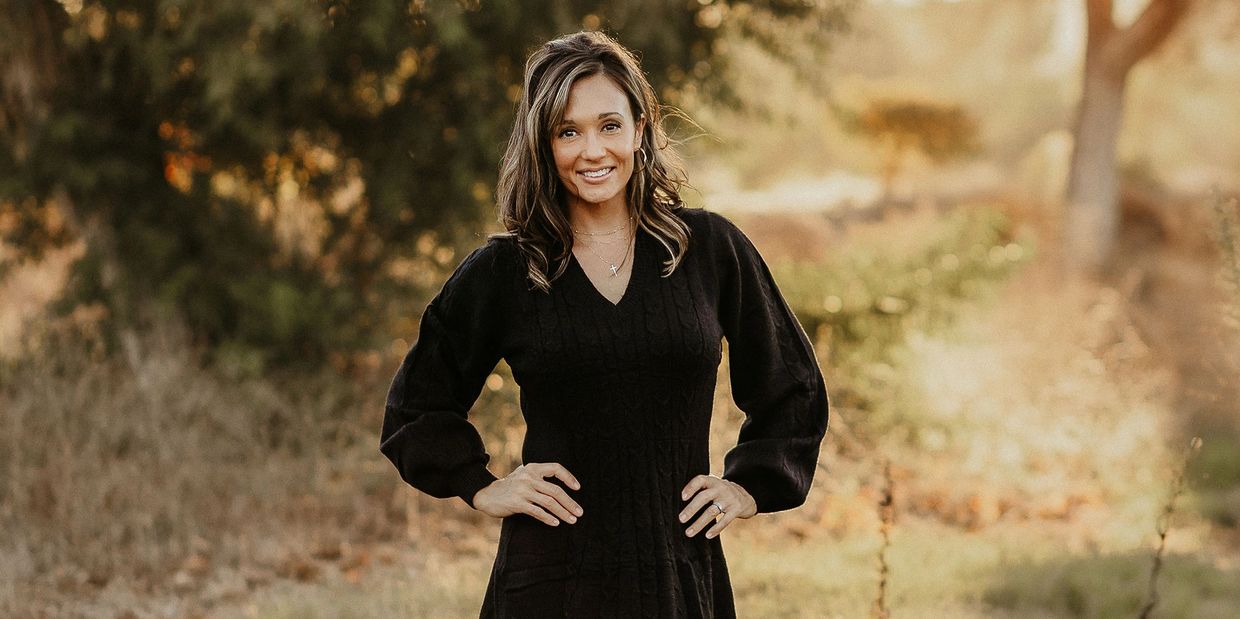A woman in a black dress smiles outdoors with hands on hips in a sunlit natural setting.