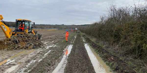 Construction workers operating machinery on a muddy rural path beside a field.