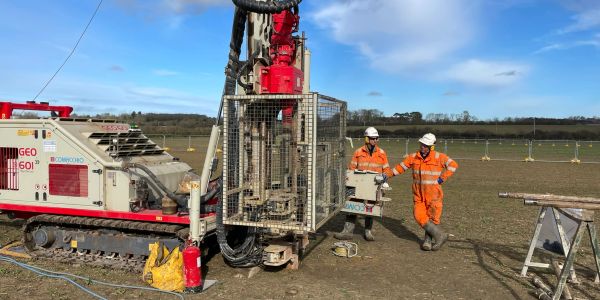 Two workers in orange gear stand by a large drilling machine in a field.