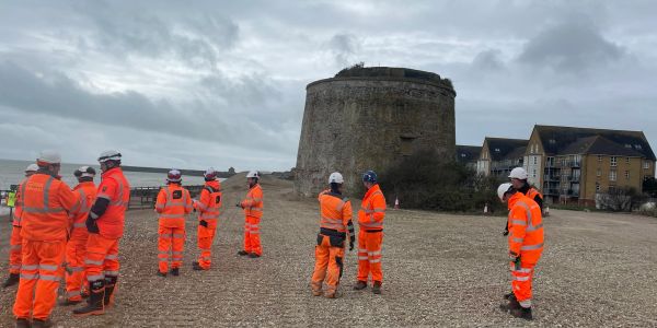 Workers in orange safety gear gathered near a historic stone tower on a cloudy day.