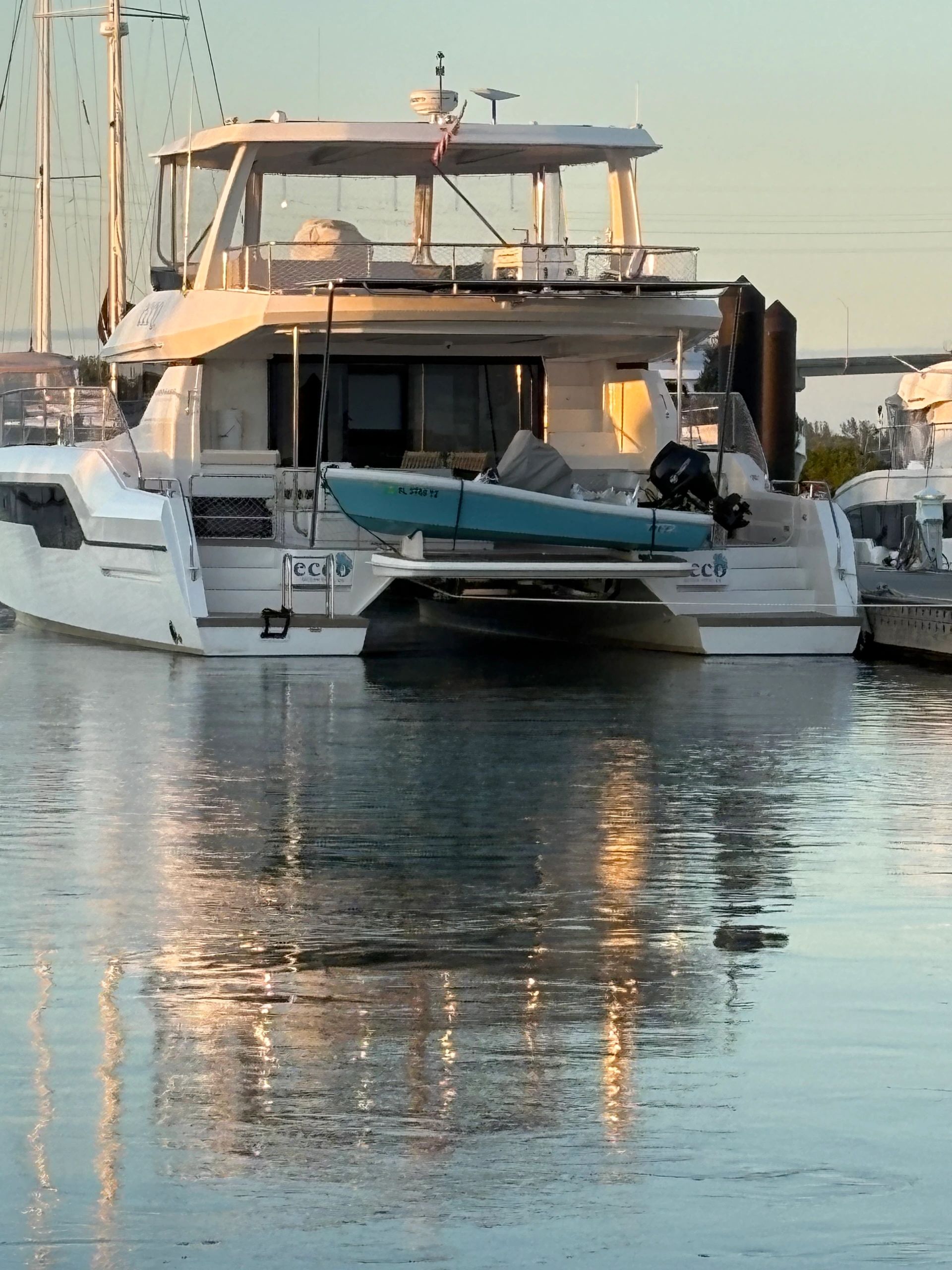 A white catamaran docked with a small boat on its platform at sunset.
