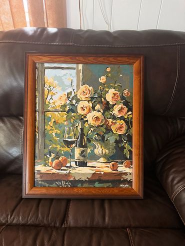 Framed painting of roses, wine, and peaches on a table near a window.