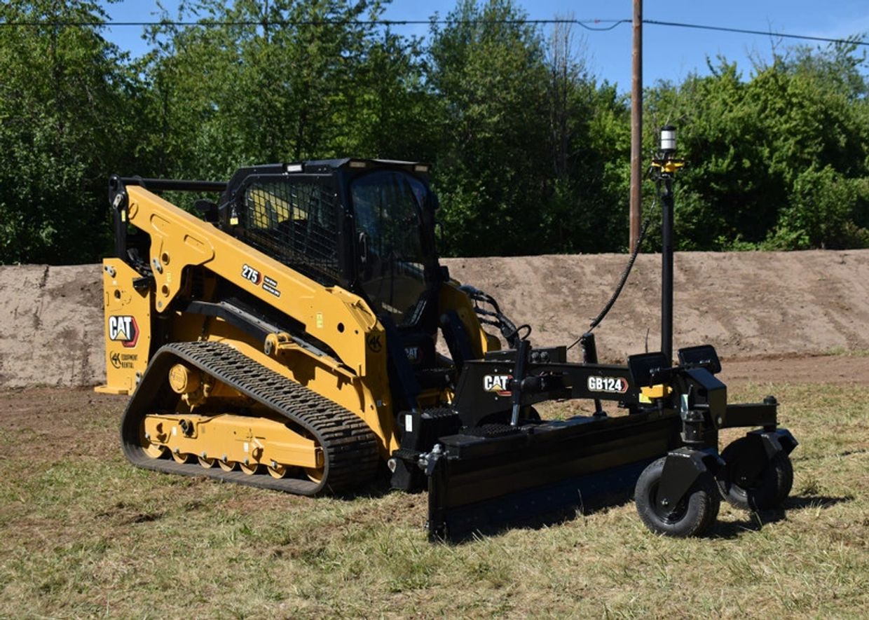 Yellow Caterpillar skid steer with a black grader attachment on grass.