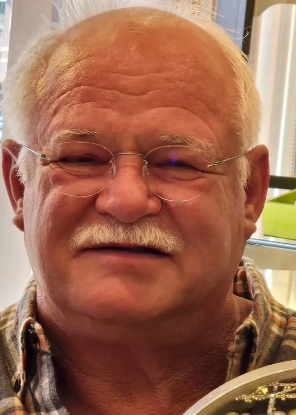 Smiling elderly man with glasses and a mustache wearing a plaid shirt.
