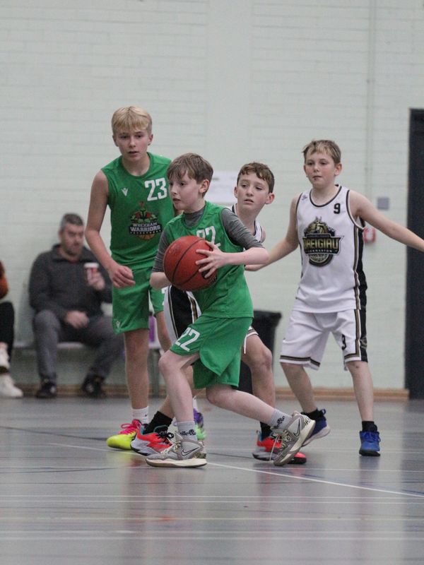 Young boys playing basketball indoors, wearing green and white jerseys. Wrexham Raiders against Dreigiau Caernarfon.