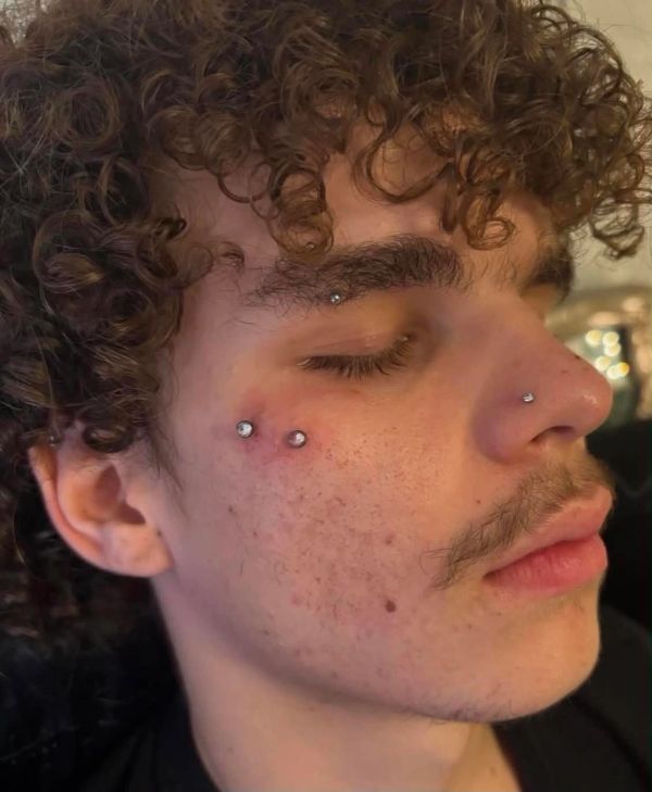 Young man with multiple facial piercings and curly hair.