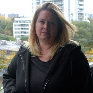 Woman with blonde hair wearing a black jacket standing outdoors in front of buildings.