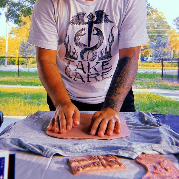 Person pressing clay on a cloth-covered table, wearing a white 'Take Care' shirt.