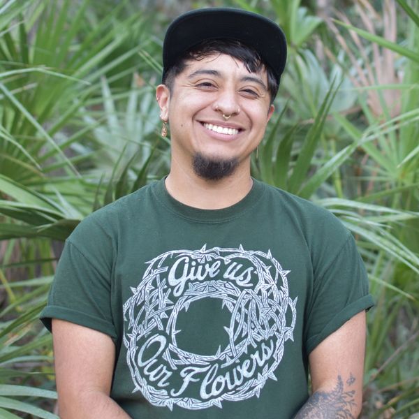 Smiling person wearing a green shirt with the text 'Give us Our Flowers' outdoors.