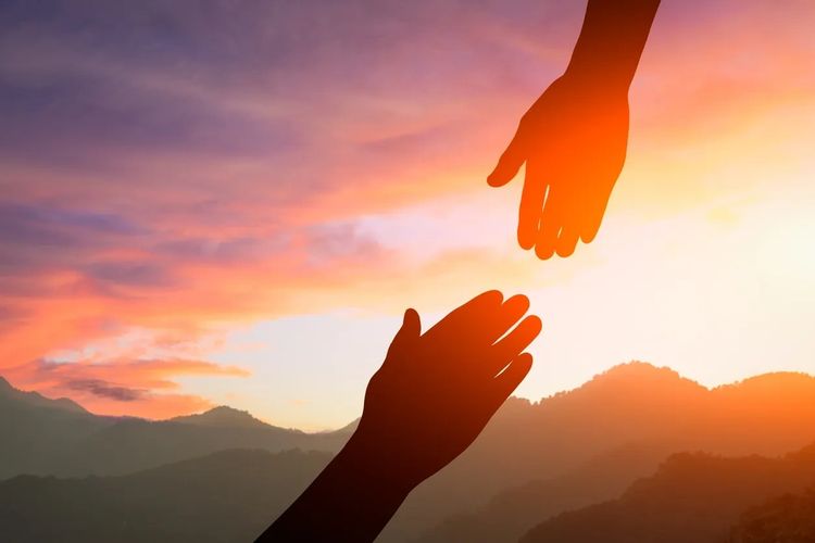 Two hands reaching out against a sunset sky over mountains.