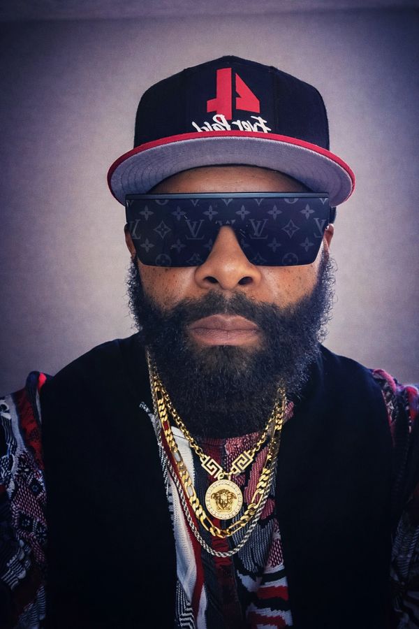 Man wearing designer sunglasses, a cap, and gold chains with a patterned shirt.
