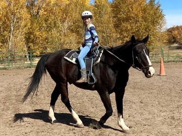 A young girl in safety gear rides a black horse in an autumn setting.