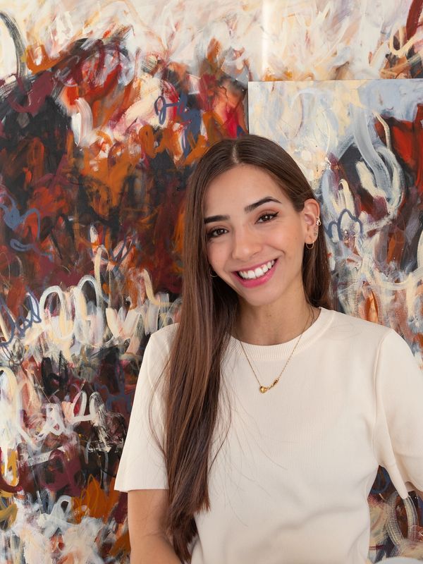 Mujer sonriente con camiseta blanca frente a pintura abstracta.