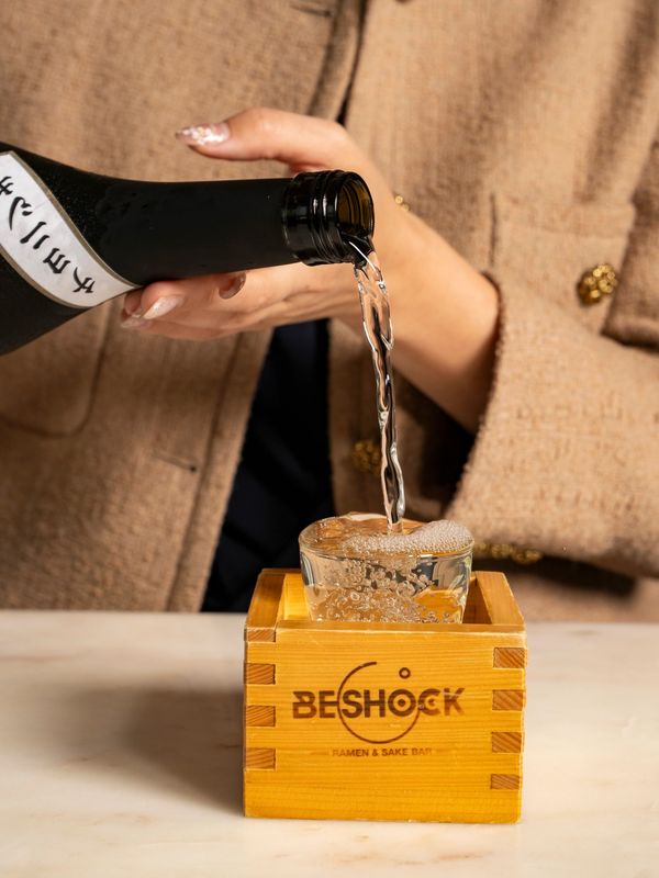 Pouring sake into a glass inside a wooden box labeled BESHOCK.