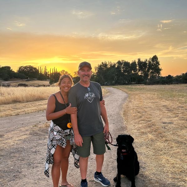 Couple with their black dog on a dirt path at sunset in a rural area.
