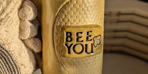Hand holding a ceramic mug with 'BEE YOU' and honeycomb pattern.
