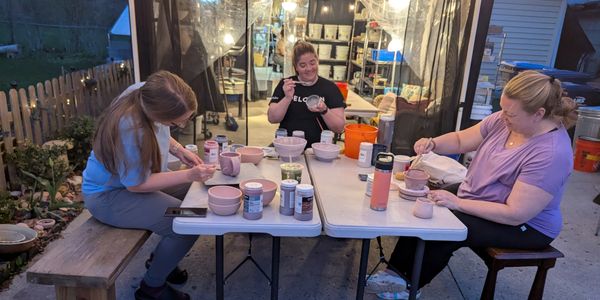 Three women painting pottery at an outdoor table in the evening.