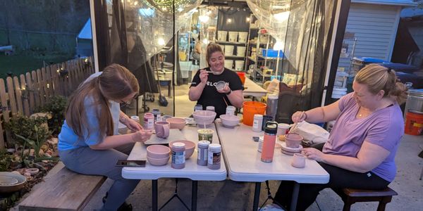 Three women painting pottery at an outdoor table in the evening.