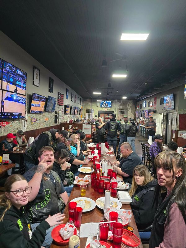 Group of bikers socializing and eating in a lively sports bar with multiple TV screens.