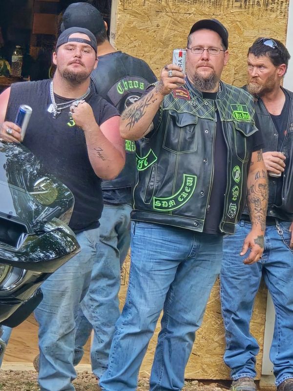 Three men in biker attire posing with drinks outdoors.