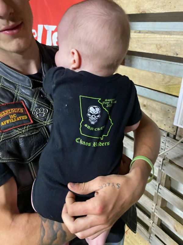 Baby in a black Chaos Riders shirt held by a tattooed person in a leather vest.