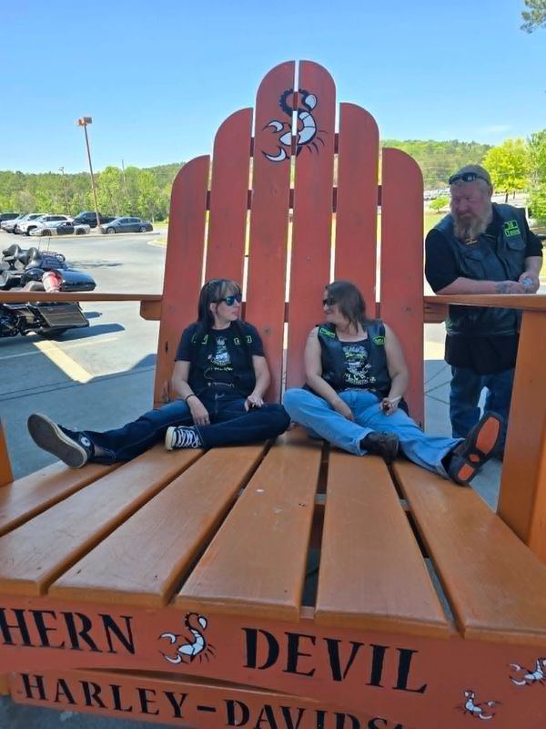 Two women and a man in biker gear relaxing on a giant wooden chair outdoors.