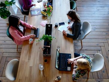 Three women working on laptops at a wooden table in a bright office.