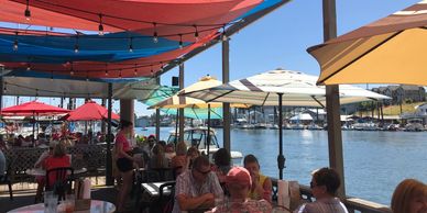 People dining outdoors under colorful umbrellas by the water on a sunny day.