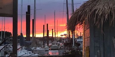 Sunset over a marina with sailboats and a thatched hut.