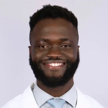 Smiling man in a white coat and tie against plain background.