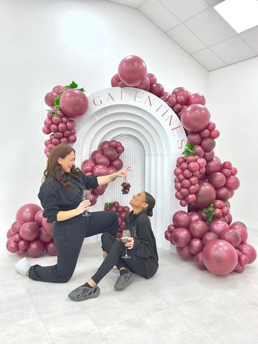 Two girls smiling holding grapes and drinking wine in front of large organic balloon display.