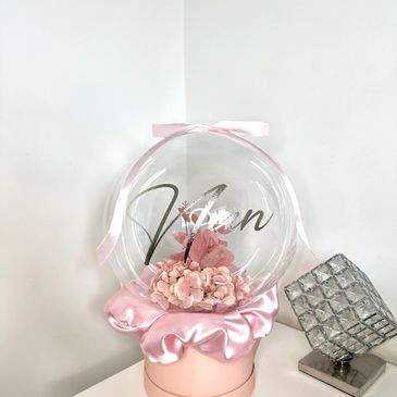 Artificial flowers inside a bobo balloon sat on top of pink satin fabric and a pink hatbox.