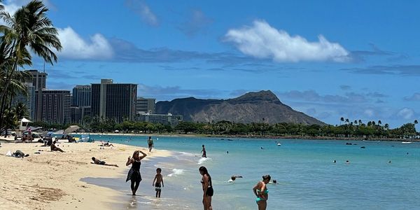 Sunny beach day with people enjoying the water and sandy shore near city buildings and mountains.