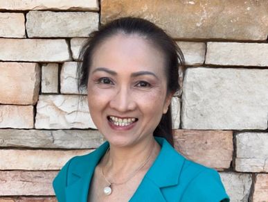 A smiling woman in a teal dress stands against a stone wall.