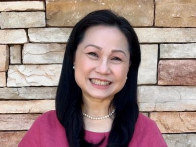 Smiling woman in a pink top and white pants stands against a stone wall.
