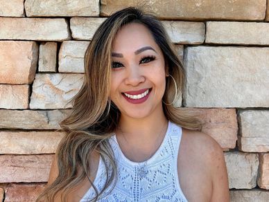 Smiling woman in white crochet top stands against a rustic stone wall.