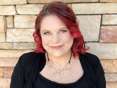 Smiling woman with red hair and a black outfit posing against a stone wall.