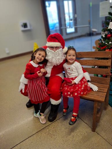 Two girls with Santa Claus in festive outfits by a Christmas tree.