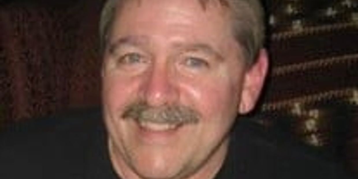Smiling man with a mustache in a dimly lit setting.