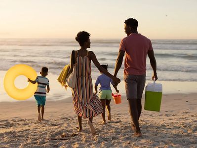 A family walks hand-in-hand along the beach at sunset, carrying beach gear
